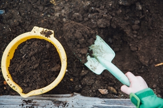 A hand holding a shovel lifting up soil to put into a yellow bucket